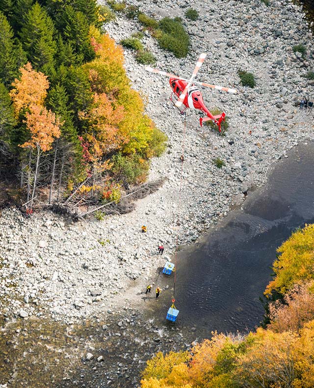 A helicopter hovers above a riverbank. Below, people work near large square containers. One container hangs from the helicopter.