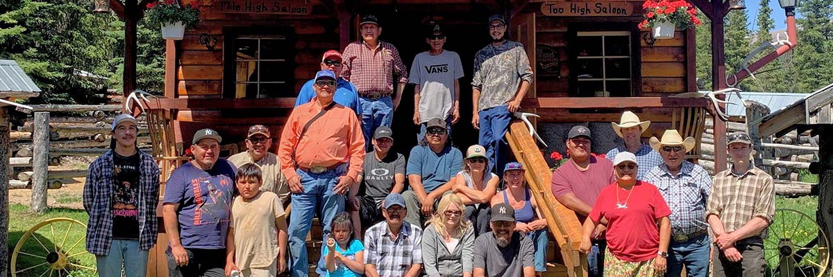 A group of 22 people of all ages, from children to Elders, pose in front of a rustic log cabin, sign posted as The Outpost at Warden Rock.