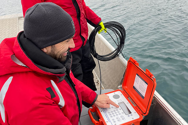 Cavan Harpur and Zachery Mielhausen on a boat in a lake, look at the display of a device attached to a cable hanging in the water.