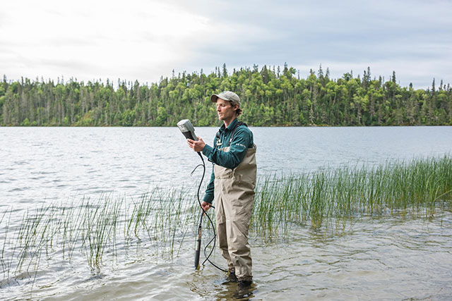 Chris Robinson stands in shallow water and short reeds. He looks at a device which connects to a probe he holds in the water.