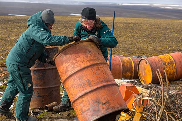 On a tundra landscape, Jane Chisholm and Angela Piercey stand up a large, rusty barrel, amid other discarded barrels and metal debris.