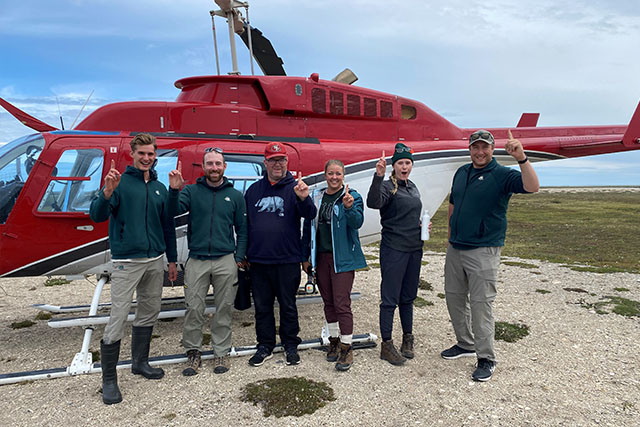 Six people pose for a group photo in front of a helicopter, each holding up one finger to signify the year one team.