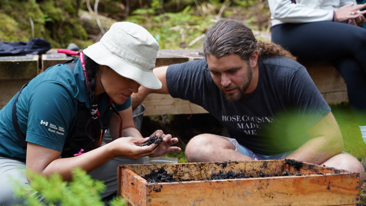 Beside a wooden box of soil, Gusdagang Mary Hart studies a dirt covered object in her hand while Jaahljuu Graham Richard leans in to look.