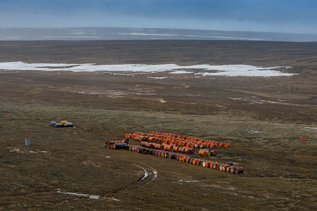 Five long rows of rusted barrels are arranged on a vast, barren tundra, with patches of snow in the distance, near a cloudy horizon.