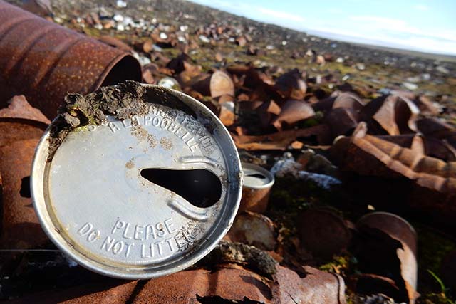 A close-up of a metal barrel top, embossed with Please Do Not Litter, resting among other barrels and metal debris, covering the landscape.