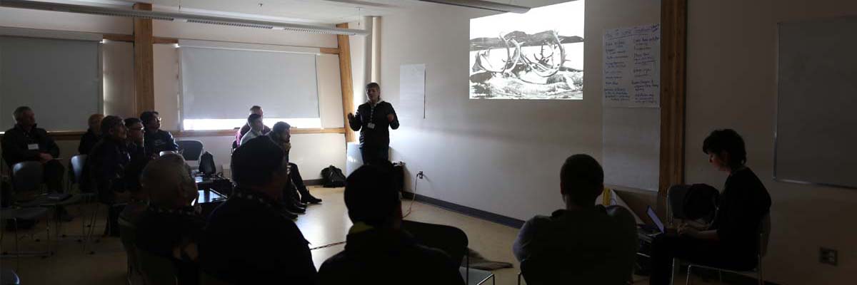 A dimly lit room with a presentation projected on a wall. Shadowed participants face the silhouette of the workshop presenter.