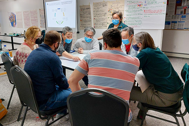 Eight engaged people sit at a table and look to where one person points on a map. Behind them, note covered chart paper hangs on the wall.