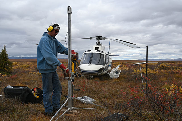 Robert Linklater sets up a tall, skinny, piece of equipment on a tripod in a tundra landscape. A helicopter is in the background.