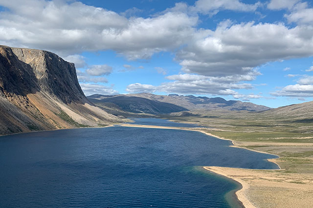 A sheer cliff descends into a calm inlet, enclosed on the other side by sloping barrens. Beyond lies rolling mountains and a sky of clouds.