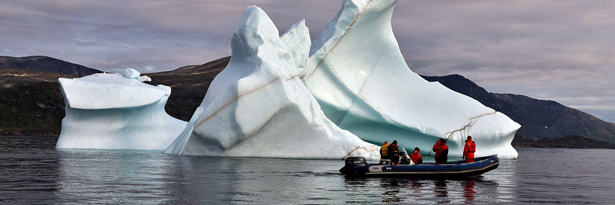 An iceberg, streaked by dark veins, juts from the water in front of a rocky island. In a small boat, five people in survival suits look on. 