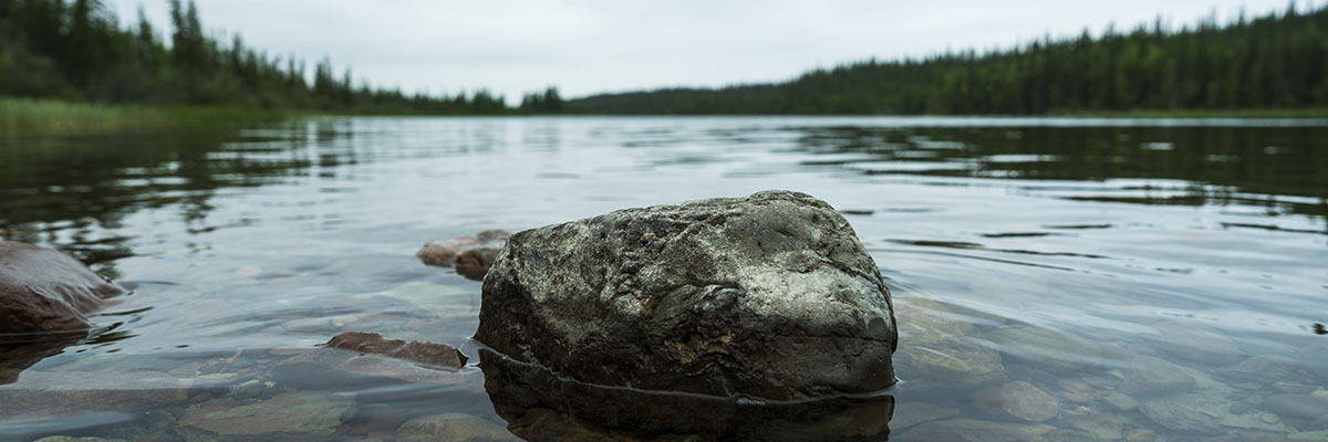 A solitary rock sits above the surface of the clear waters of a calm lake with a rocky bottom. Coniferous forests line the shore.