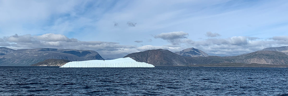 An immense flat iceberg, showing a furrowed edge, protrudes from the ocean in front of an undulating, mountain landscape, and expansive sky.