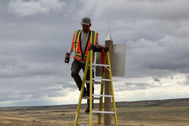 Chris Reed in a hi-vis vest, stands on a ladder, installing a pointed cone on the top of a signpost in the grasslands.