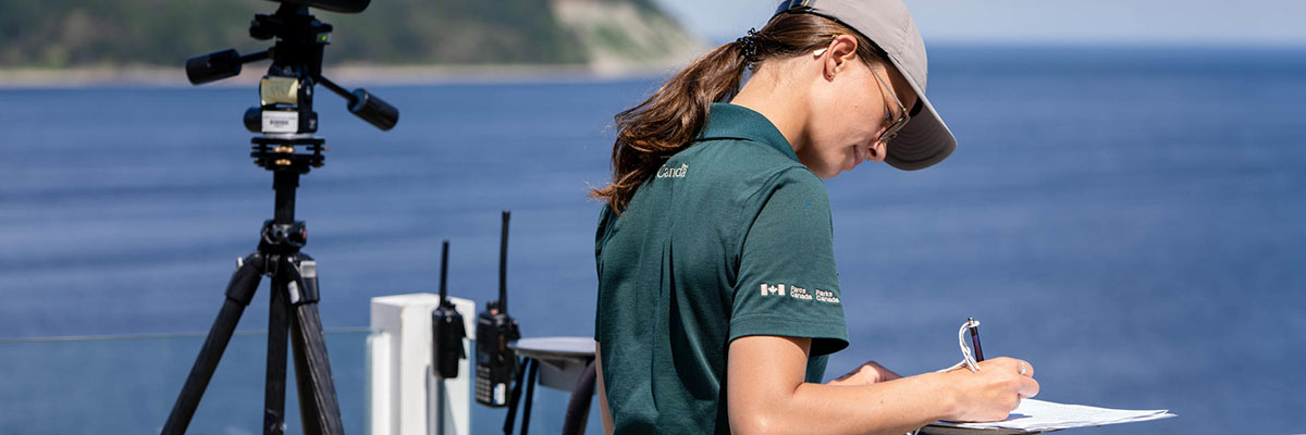 Catherine Blanchette in uniform, stands on a viewing platform, writing on a clipboard, next to a telescope, radios and binoculars.