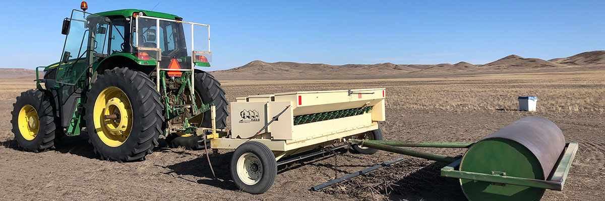 A tractor pulls seeding equipment and a barrel shaped roller across an open grassy plain, under a clear sky.